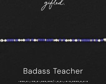 May include: A beaded bracelet featuring alternating dark blue and metallic silver cylindrical beads arranged in a linear pattern. The text gifted. appears above the bracelet, while Badass Teacher and Morse code dots and dashes appear below.