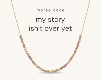 May include: A gold chain necklace featuring a pattern of small metallic gold beads and mauve-coloured cylindrical beads arranged in a Morse code sequence. The text "morse code" and "my story isn't over yet" is printed above the necklace.