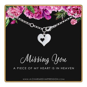 May include: A silver necklace with a heart pendant featuring a smaller heart cutout. The necklace is displayed on a black card with the words "Missing You" and "A Piece of My Heart is in Heaven". Pink flowers are in the background.