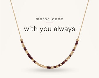 May include: A delicate gold chain necklace featuring a sequence of small metallic gold and dark red beads arranged in a Morse code pattern. The text "morse code" and "with you always" is printed in black above the necklace on a plain background.