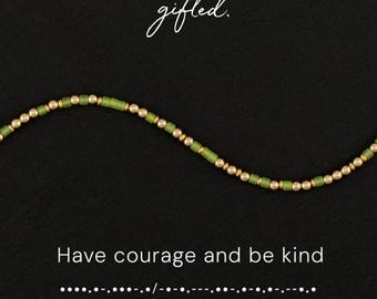 May include: A beaded necklace featuring alternating metallic gold spheres and matte olive green cylindrical beads. The text gifted. appears at the top, while the phrase Have courage and be kind and Morse code dots and dashes appear below.