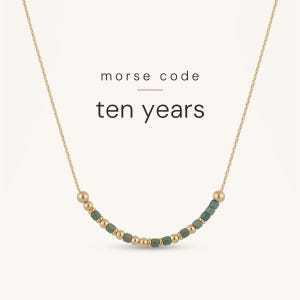 May include: A gold chain necklace featuring a central section of alternating small gold-toned spherical beads and dark teal cylindrical beads arranged in a Morse code pattern. The text "morse code" and "ten years" is printed above the necklace.