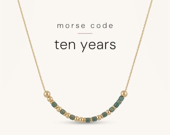 May include: A gold chain necklace featuring a central segment of alternating small gold-toned spherical beads and cylindrical sage green beads. The text "morse code" and "ten years" is printed in black above the beaded section of the necklace.