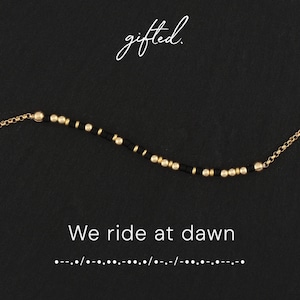 Könnte beinhalten: Ein goldenes Kettenarmband mit einer Abfolge kleiner schwarzer und goldener Perlen in einem Morsecode-Muster. Der Text gifted. erscheint über dem Armband, während der Text We ride at dawn und der zugehörige Morsecode darunter stehen.