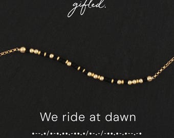 May include: A delicate bracelet featuring alternating matte black cylindrical beads and small gold-toned spherical beads. The text gifted. appears above the bracelet, while the text We ride at dawn and Morse code dots and dashes appear below.
