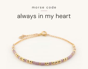 May include: A gold chain bracelet featuring a pattern of small, round gold beads and cylindrical mauve beads arranged in Morse code. The text above the bracelet reads morse code always in my heart in black lettering on a plain background.