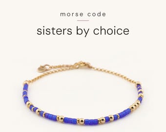 May include: A gold chain bracelet featuring blue and gold beads arranged in a Morse code pattern. The text above the bracelet reads morse code sisters by choice. The bracelet includes a delicate gold chain with a small circular charm.