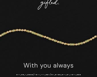 May include: A beaded necklace featuring alternating matte sage green cylindrical beads and small metallic gold spherical beads. The text gifted. appears at the top, while the phrase With you always and Morse code are printed below.