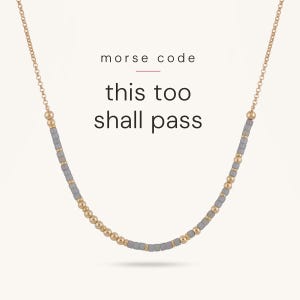 May include: A gold chain necklace featuring a pattern of small round gold beads and grey cylindrical beads arranged in Morse code. The text "morse code" and "this too shall pass" is printed in black above the necklace.