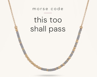 May include: A gold chain necklace featuring a pattern of small round gold beads and grey cylindrical beads arranged in Morse code. The text "morse code" and "this too shall pass" is printed in black above the necklace.