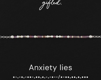 May include: A delicate bracelet featuring a sequence of dark purple and metallic silver beads arranged in a Morse code pattern. The text "gifted." appears above the bracelet, while "Anxiety lies" and the corresponding Morse code are below.