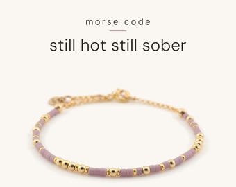 May include: A delicate bracelet featuring alternating small gold-tone metallic beads and matte lavender-coloured cylindrical beads. The text above the bracelet reads morse code still hot still sober in black font with a thin horizontal line.