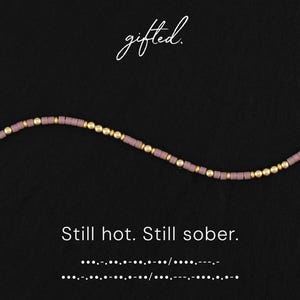 May include: A necklace with a dark background. The necklace is made of small, rectangular, mauve-colored beads and gold-colored round beads. The text "gifted" is written in cursive at the top. The text "Still hot. Still sober." is written below the necklace.