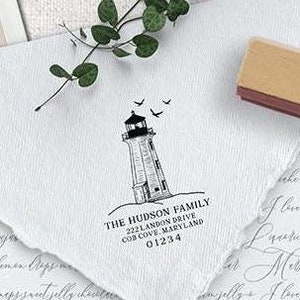 May include: A white envelope with a hand-drawn lighthouse illustration and the text "THE HUDSON FAMILY 222 LAMBON DRIVE COS COVE MARYLAND 01234" printed on it. The envelope is on a white surface with a wooden stamp and a green plant.