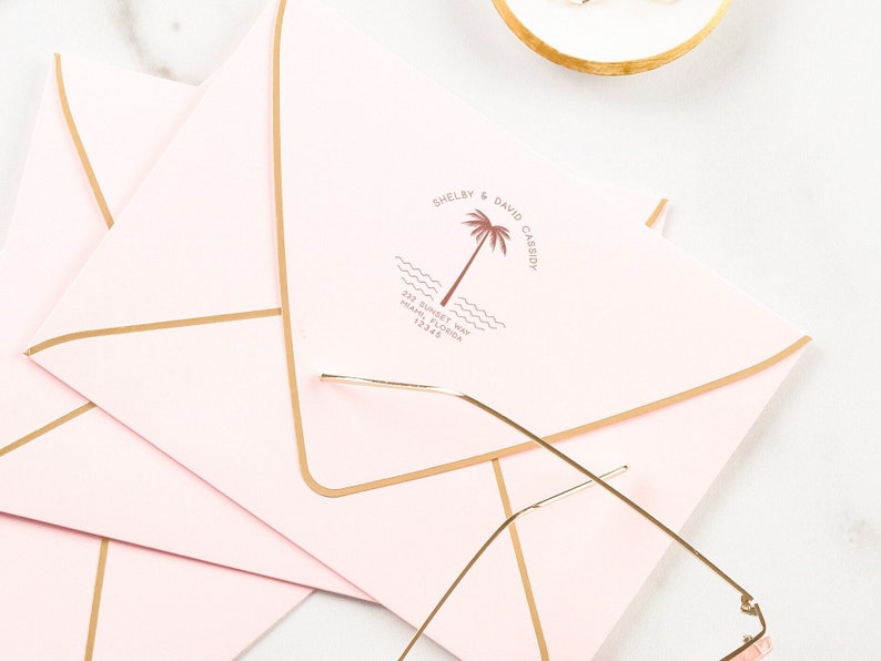 May include: Pink envelopes with gold trim and a custom return address stamp featuring a palm tree and the text "Shelby & David Cassidy" and "22nd June 2024 Palm Beach, FL 12:30 PM".