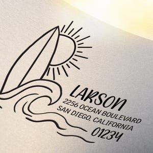 May include: Black and white line art design featuring a surfboard, sun, and waves. The name "Larson" and address details are printed below the graphic. The design is on a light-colored background.