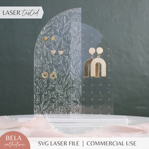 Könnte beinhalten: Ein transparenter Acryl-Ohrringständer mit einem Blumenmuster auf der einen Seite und einem Lochraster auf der anderen. Der Ständer ist halbkreisförmig und hat eine weiße Basis. Der Text "LASER tested" ist in der oberen linken Ecke des Bildes sichtbar.
