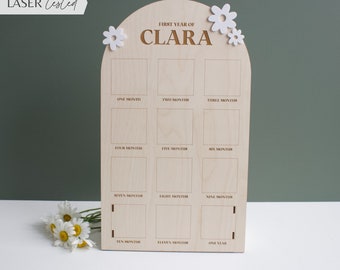 Puede incluir: Un gráfico de crecimiento de madera con un arco blanco y detalles de flores blancas. El gráfico está etiquetado como "Primer año de Clara" y tiene 12 cuadrados para marcar el crecimiento de un niño cada mes.