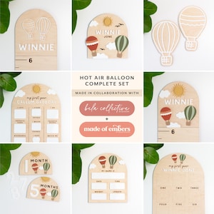 May include: A set of wooden hot air balloon themed baby milestone markers. The set includes a growth chart, a first year chart, a monthly milestone chart, and a name sign. The markers are made of wood and painted in a light wood colour. The balloons are painted in various colours, including red, green, and white. The set is made in collaboration with Bela Collective and Made of Embers.