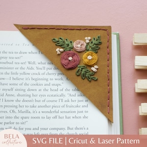 May include: A brown leather bookmark with a floral embroidery design in shades of pink, yellow, and green. The bookmark is folded into a triangle shape and is placed on top of a book. The text "BELA collective" and "SVG FILE | Cricut & Laser Pattern" is visible at the bottom of the image.