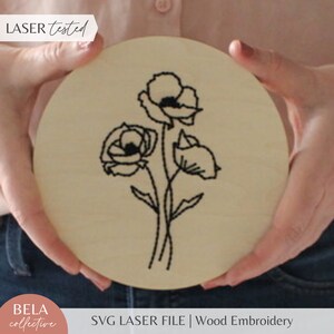 Könnte beinhalten: Runde Holzplakette in hellem Farbton mit schwarzem Blumenstickerei-Design. Die Plakette wird von Händen gehalten und trägt die Aufschrift "LASER tested" und "SVG LASER FILE | Wood Embroidery".