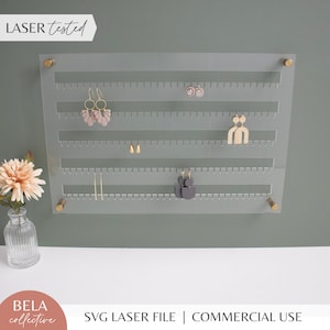 May include: A clear acrylic earring holder with gold accents and rows of holes for hanging earrings. The holder is mounted on a green wall. "LASER tested" is written in white on a black rectangle at the top of the image.  "BELA collective" is written in white on a pink circle at the bottom of the image.  "SVG LASER FILE | COMMERCIAL USE" is written in black at the bottom of the image.