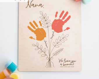 May include: A wooden sign with the text "Nana" and two handprints in orange and red paint. The handprints are arranged to look like flowers with stems and leaves. The text "We love you a bunch!" is below the handprints. The text "Mother's Day 2023" is below the previous text.