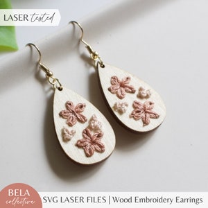May include: A pair of wooden teardrop-shaped earrings with gold hooks. The earrings are decorated with embroidered flowers in shades of pink and beige.