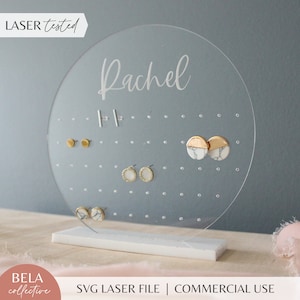 Peut inclure: Un porte-boucles d'oreilles rond en acrylique transparent avec une base blanche. Le porte-boucles d'oreilles porte une inscription personnalisée qui dit "Rachel". Le porte-boucles d'oreilles a de multiples trous pour les boucles d'oreilles. Il y a plusieurs paires de boucles d'oreilles sur le porte-boucles d'oreilles, y compris des boucles d'oreilles en or et en marbre.