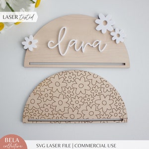 Puede incluir: Un letrero de madera en forma de semicírculo con el nombre "Clara" escrito en cursiva blanca. El letrero tiene un pequeño estante y está decorado con flores blancas. El letrero está cortado con láser y probado.