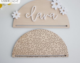 May include: A wooden half-circle sign with the name "Clara" written in white script. The sign has a small shelf and is decorated with white flowers. The sign is laser cut and tested.