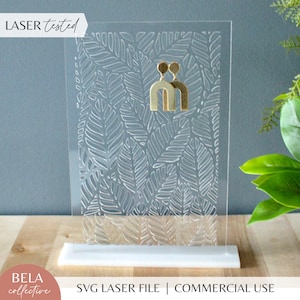 May include: A clear acrylic earring display stand with a laser-cut design of leaves. The stand has a white base and features the text "LASER tested" and "SVG LASER FILE | COMMERCIAL USE".