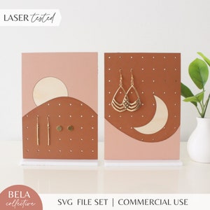 May include: Two wooden earring display stands with a pink and brown color scheme. One stand features a crescent moon and the other features a sun and hills. Both stands have holes for displaying earrings.