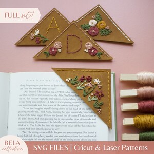 May include: A set of three brown felt triangle bookmarks with embroidered floral designs and the letters A, D, and a flower. The bookmarks are on a pink surface with spools of embroidery floss.