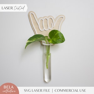 May include: A wooden hand-shaped cutout with a white outline, designed to hold a glass test tube vase with a green plant. The text "LASER tested" is visible in the top left corner of the image.