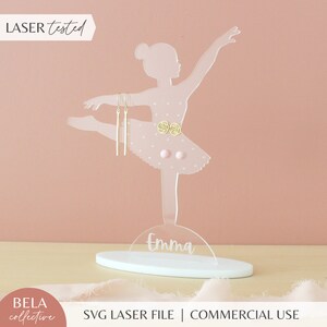 Könnte beinhalten: Klare Acryl-Ballerina-Ohrringhalterung mit weißem Sockel. Die Ballerina ist in einer Tanzpose mit einem rosa Tutu und einem goldenen Gürtel. Der Sockel trägt den Namen "Emma".