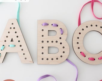 May include: Wooden letters A, B, and C with holes for lacing. The letters are painted in natural wood color.  The letters are being laced with colorful ribbons.  The text "LASER tested" and "SVG LASER FILES | Wood Lacing" are visible in the image.