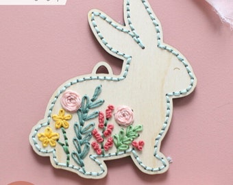 May include: A wooden bunny shaped ornament with a floral embroidery design. The bunny is white with a light blue outline and the embroidery features pink, yellow, green, and red flowers.