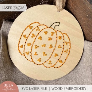 May include: A round wooden ornament with a stitched orange pumpkin design. The pumpkin has a dotted pattern and the text "LASER tested" is visible in the top left corner.