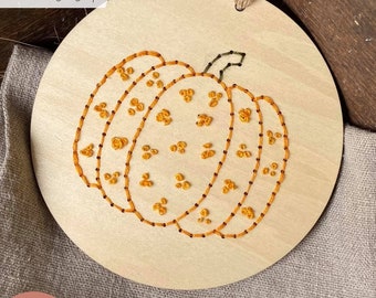 May include: A wooden round ornament with a stitched orange pumpkin design. The pumpkin has a dotted pattern and the text "LASER tested" is visible in the top left corner.