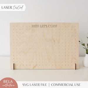 May include: A wooden pegboard with holes for displaying earrings or other small items. The pegboard is light brown and has the text "HEY! LET'S CLAY!" printed on it.
