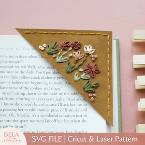 May include: A brown leather bookmark with a floral embroidery design. The embroidery features pink, white, and green flowers. The bookmark is folded over the corner of a book.