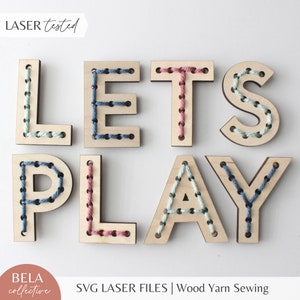 Puede incluir: Letras de madera que deletrean "LET'S PLAY" con hilo cosido a través de agujeros preperforados. Las letras están pintadas en tonos de madera natural y presentan hilo de colores en azul, rosa y blanco.