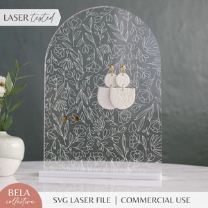 May include: Clear acrylic earring display stand with a floral design. The stand features a white base and an arched top with etched flowers and leaves. Two pairs of white earrings with gold accents are displayed. The text "LASER tested" and "SVG LASER FILE | COMMERCIAL USE" are visible.