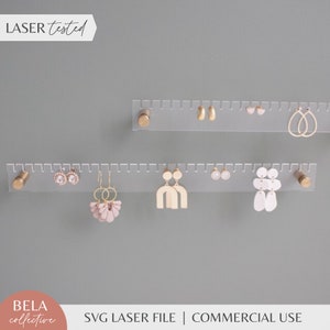 May include: Two clear acrylic earring organizers with gold metal end caps. The organizers have a ruler-like design with evenly spaced holes for hanging earrings. The top organizer has gold, white, and pink earrings. The bottom organizer has gold, white, and pink earrings. The text "LASER tested" is in the top left corner of the image. The text "BELA collective" is in the bottom left corner of the image. The text "SVG LASER FILE | COMMERCIAL USE" is in the bottom center of the image.