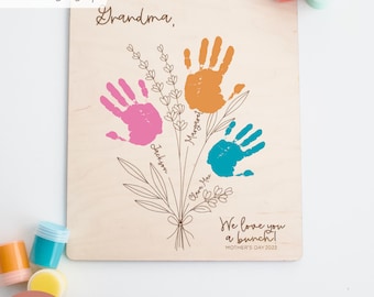 May include: A wooden sign with a handprint flower bouquet design. The sign says "Grandma, We love you a bunch!" and "Mother's Day 2023".