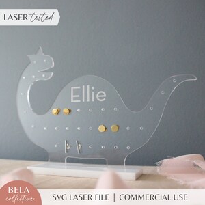May include: Clear acrylic dinosaur earring holder with the name "Ellie" engraved on the side. The holder has multiple holes for earrings and is standing on a white base.