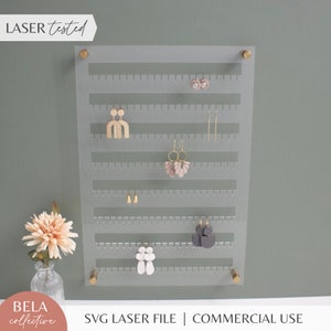 Peut inclure: Un porte-boucles d'oreilles en acrylique transparent avec 7 rangées de trous pour accrocher les boucles d'oreilles. Le support est doté d'accents en métal doré et est monté sur un mur vert. Le texte "LASER tested" est visible dans le coin supérieur gauche de l'image. Le texte "SVG LASER FILE | COMMERCIAL USE" est visible en bas de l'image.