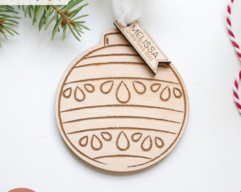 Puede incluir: Un adorno navideño de madera con un diseño grabado con láser. El adorno es redondo con líneas horizontales y formas de lágrima. Una pequeña etiqueta dice "MELISSA CHRISTMAS 2023". También se ven las palabras "LASER tested".
