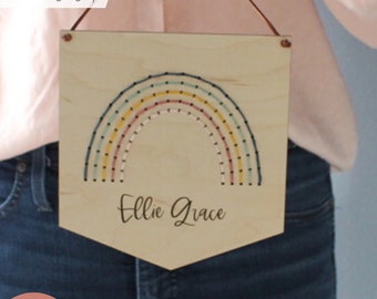 Puede incluir: Un letrero de madera con un diseño de arcoíris y el nombre "Ellie Grace" escrito debajo. El arcoíris está hecho de hilo de colores y tiene puntos negros a lo largo de los bordes. El letrero está colgado de un cordón marrón.